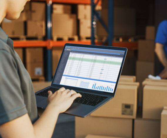 Female Manager Using Laptop Computer To Check Inventory. In the Background Warehouse Retail Center with Cardboard boxes, e-Commerce Online Orders, Food, Medicine, Products Supply. Over the Shoulder