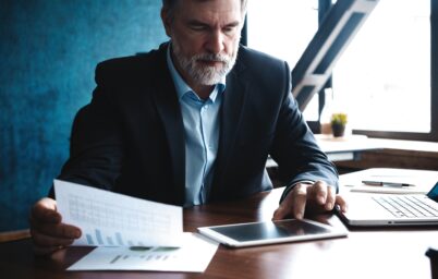 Serious mature financial advisor sitting at the table and checking financial report using digital tablet at office.